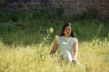 Beyaz elbiseli, genç, güzel, esmer kadın, kameraya bakıyor, rahat ve sakin, rüzgarlı bir günde uzun otların arasında oturuyor. Güzellik, moda, moda, gençlik kavramı.