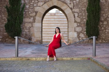 Genç, güzel, esmer, güzel kırmızı parti elbisesi içinde gülümseyen, mutlu ve komik poz veren bir kadın sokakta büyük bir zincirde oturuyor. Güzellik, moda, moda, gençlik kavramı.