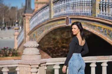 Genç, güzel, siyah uzun kollu tişörtlü ve kot pantolonlu bir kadın, arkası dönük, kameraya bakıyor, Seville 'de İspanya meydanında bir korkuluğa yaslanıyor..