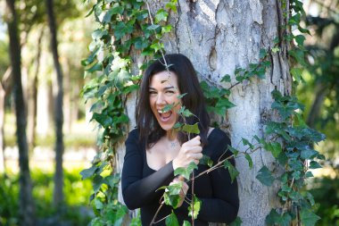 Genç, güzel, esmer bir kadın, siyah tişört ve kot pantolon, bir ağacın gövdesine yaslanmış, sarmaşıkla komik oynuyor, doğayla bağlantılı. Güzellik kavramı, kadınlık, doğa, bitkiler, ağaçlar.