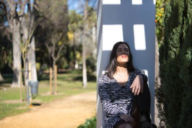 Genç, güzel, siyah saçlı bir kadın, zebra elbisesi içinde, gözleri kapalı, oturuyor ve sırtı bir sütunun üzerinde dinleniyor, güneş ışınlarını yüzüne alıyor. Kavramsal ışıklar, gölgeler.