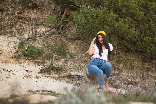 Genç, güzel bir kadın, siyah saçlı, beyaz gömlekli, kot pantolonlu ve sarı eşarplı, gülümseyen ve mutlu bir şekilde kameraya bakan, bir ağacın kökünde oturan. Güzellik, moda, plaj, doğa kavramları.
