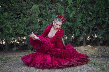 Genç, güzel, kırmızı flamenko elbiseli sarışın kadın, saçında çiçekler ve altın mücevherler, fonda yeşil bitkilerle yerde otururken flamenko pozları veriyor. Kavram dansı, sanat.