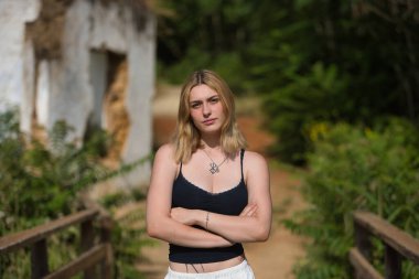 Genç, güzel, siyah tişörtlü, beyaz pantolonlu, özgür ve güçlü, kolları çapraz bir şekilde açık hava parkında kameraya bakan sarışın bir kadın. Güzelliği kavra, gençliği, milenyumu.