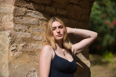 Genç, güzel, sarışın, siyah gömlekli, masum ve hassas bir kadının portresi. Kameraya bakıyor, tuğla kemere yaslanıyor, açık hava parkında güneş ışınları alıyor. Güzellik kavramı, gençlik.