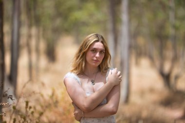 Genç, güzel, sarışın bir kadın, çiçekli bir elbise giyiyor, kendini kucaklıyor, kendini seviyor ve bir koruma işaretiyle, açık hava parkında. Güzellik kavramı, gençlik, milenyum.