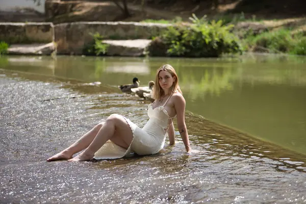 Beyaz elbiseli, genç, güzel, sarışın bir kadın, rahat ve sakin, açık hava parkında ördekleri olan bir gölde küçük bir şelalede oturuyor. Güzelliği kavra, gençliği, milenyumu.