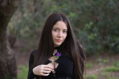 Genç, koyu renk saçlı, soluk tenli, siyah bir elbise giyen, elinde mor bir çiçekle baharın gelişini kutlayan güzel bir kadının portresi..