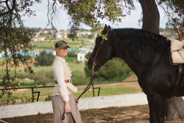 Genç bayan binici, siyah İspanyol safkan atının yanında poz veriyor, dizginleri elinde tutuyor. Konsept at biniciliği, at biniciliği, binicilik, binicilik.