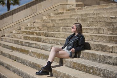 Genç, güzel, sarışın, deri ceketli, yılan desenli mini etekli, çizmeli ve güneş gözlüklü, rahat ve sakin güneşlenen, merdivenlerde oturan. İsyankar, boyun eğmez, isyancı konsept.