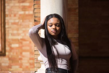 Genç, güzel, Afrikalı bir kadın, süveteri ve deri şortu var, gözleri kapalı ve eli başında, güneşleniyor, Seville 'de İspanya meydanında bir sütuna yaslanıyor. Moda konsepti, güzellik, moda.