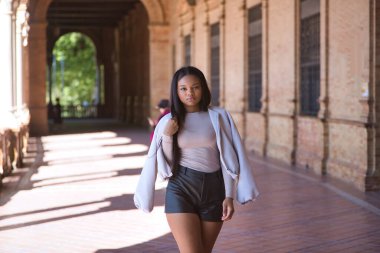 Genç, güzel, Afrikalı bir kadın, ceketli ve deri şortlu, İspanya meydanında poz veriyor ve modellik yapıyor. Moda konsepti, güzellik, moda.