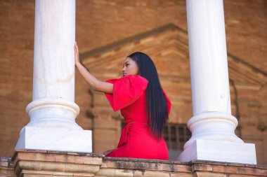 Genç, güzel, kırmızı elbiseli Afrikalı bir kadın. İspanya 'nın Sevilla meydanındaki iki mermer sütunun arasında sonsuzluğa bakıyor. Moda konsepti, güzellik, moda.