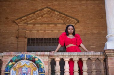 Genç, güzel, kırmızı elbiseli Afrikalı bir kadın, Seville 'de İspanya meydanındaki korkuluklara yaslanan kameraya ciddi bir jest yapıyor. Moda konsepti, güzellik, moda.