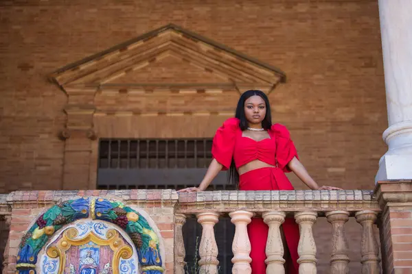 Genç, güzel, kırmızı elbiseli Afrikalı bir kadın, Seville 'de İspanya meydanındaki korkuluklara yaslanan kameraya ciddi bir jest yapıyor. Moda konsepti, güzellik, moda.