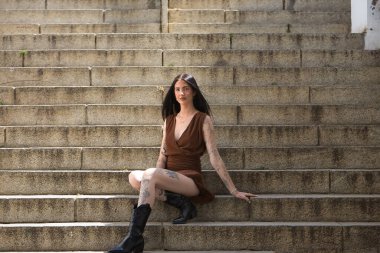 Genç bir kadın, esmer, uzun boylu, zayıf ve çok dövmeli, kahverengi elbiseli ve çizmeli, tatlı, masum ve yalnız, merdivenlerde oturuyor. Gençlik kavramı, güzellik, asilik, milenyum.