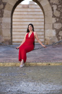 Genç, güzel, esmer, güzel kırmızı parti elbisesi içinde gülümseyen, mutlu ve komik poz veren bir kadın sokakta büyük bir zincirde oturuyor. Güzellik, moda, moda, gençlik kavramları. Dikey konum.