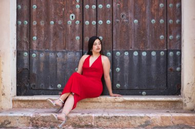 Genç, güzel, esmer bir kadın, güzel kırmızı bir parti elbisesi ve topuklu ayakkabıları içinde, rahat ve sakin, bazı basamaklarda oturuyor ve eski bir ahşap kapıya yaslanıyor. Güzellik, moda, moda, gençlik kavramı.