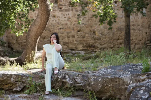 Genç, güzel, beyaz elbiseli ve topuklu ayakkabılı esmer kadın, elleri çenesinde, düşünceli ve konsantre, büyük bir kayanın üzerinde oturuyor. Güzellik, moda, moda, gençlik kavramı.