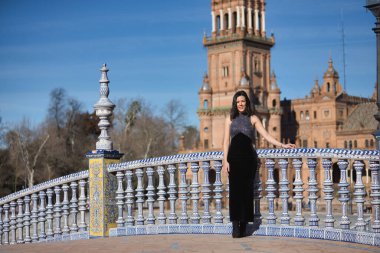 Genç, güzel, esmer, süslü siyah elbiseli bir kadın kameraya bakıyor, Seville 'de İspanya meydanındaki bir köprüde fayanslı bir korkuluğa yaslanıyor. Güzelliği, kadınlığı, turizmi kavrayın.