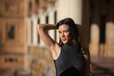 Süslü, siyah elbiseli esmer bir kadının portresi, rahat ve sakin bir şekilde kameraya bakıyor, mermer bir sütuna yaslanıp oturuyor. Güzelliği, modayı, modayı, kadınlığı kavra.