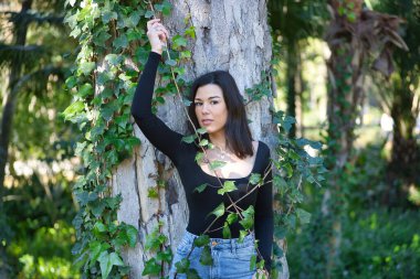 Siyah tişört ve kot pantolon giymiş, bir ağacın gövdesine yaslanmış ve doğayla bağlantılı, sarmaşığa dolanmış genç, güzel, koyu renk saçlı bir kadın. Güzellik kavramı, kadınlık, doğa, bitkiler, ağaçlar.