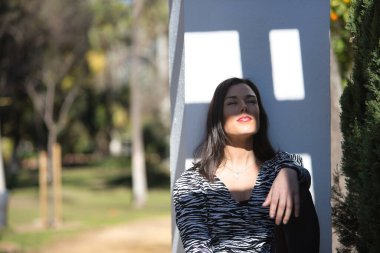 Genç, güzel, siyah saçlı bir kadın, zebra elbisesi içinde, gözleri kapalı, oturuyor ve sırtı bir sütunun üzerinde dinleniyor, güneş ışınlarını yüzüne alıyor. Kavramsal ışıklar, gölgeler.