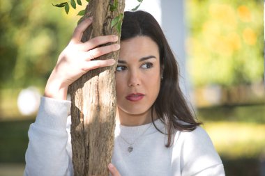 Beyaz elbiseli, genç, güzel, koyu renk saçlı bir kadının portresi, tatlı ve rüya gibi bakışlı, bir sarmaşığın gövdesine yapışan. Güzellik, moda, eğilim, kadınlık, doğa, bitkiler, ağaçlar.