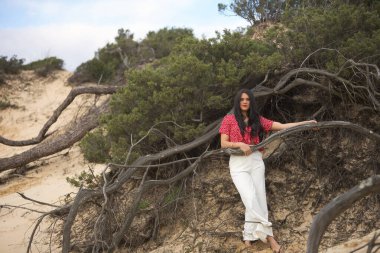 Genç, güzel, esmer, kırmızı gömlekli ve beyaz pantolonlu bir kadın, kameraya bakıyor rahat ve sakin, kumun içine düşmüş bir ağacın köklerine tutunuyor. Güzelliği kavra, rahatla, doğa, sakinlik.