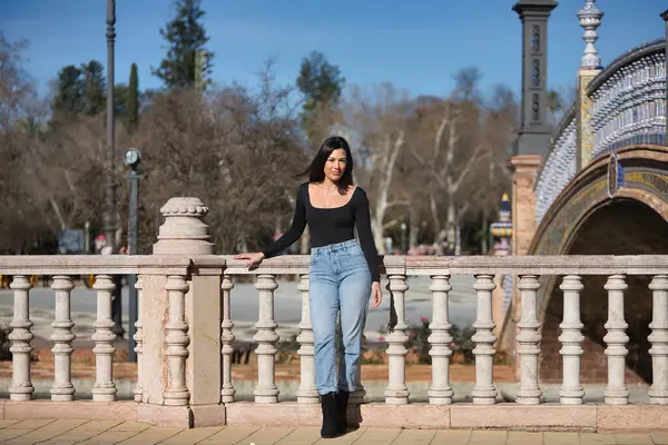 Genç, güzel, esmer bir kadın, siyah uzun kollu tişört ve kot pantolon giyiyor, saçları rüzgarda uçuşuyor, Seville 'de İspanya meydanında bir korkuluğa yaslanmış poz veriyor. Güzellik kavramı, kadınlık.