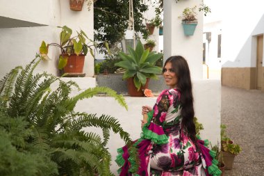 Genç, güzel, esmer, flamenko dansçısı, güzel elbisesini gösteren, arkası dönük bir sandalyede oturan, güzel ve tipik Endülüs avlusunda bir kuyunun yanında.