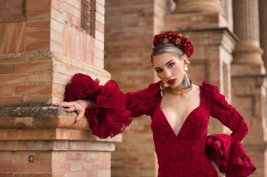 Genç, güzel, kırmızı flamenko elbiseli sarışın kadın, saçında çiçekler ve altın mücevherler, Seville 'de İspanya meydanında tuğla bir duvara yaslanmış poz veriyor. Dans konsepti, sanat, tipik İspanyol..