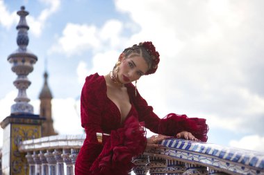 Genç, güzel, sarışın bir kadının portresi, kırmızı flamenko elbiseli, saçlarında çiçekler ve altın mücevherlerle, İspanya 'nın Seville meydanındaki güzel bir köprüde flamenko pozları veriyor. Dans konsepti.