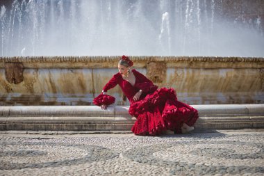 Genç, güzel, sarışın, kırmızı flamenko elbiseli, saçında çiçekler ve altın mücevherlerle, İspanya 'nın Seville meydanındaki güzel bir çeşmede flamenko pozları veriyor. Dans konsepti, sanat.