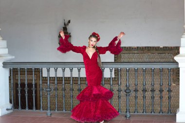 Genç, güzel, sarışın, kırmızı flamenko elbiseli, saçında çiçekler ve altın mücevherlerle, Seville 'de İspanya meydanında metal bir korkuluğa yaslanmış flamenko pozları veriyor. Dans konsepti, sanat.