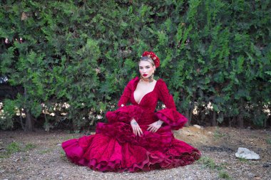 Genç, güzel, kırmızı flamenko elbiseli sarışın kadın, saçında çiçekler ve altın mücevherler, fonda yeşil bitkilerle yerde otururken flamenko pozları veriyor. Kavram dansı, sanat.