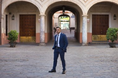 Genç, esmer, İspanyol, takım elbiseli ve kravatlı modern yönetici, elleri cebinde yalnız bir sokakta poz veriyor. Konsept iş, şirket, lider.