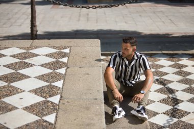 Genç, esmer, İspanyol, modern adam, siyah beyaz çizgili örgü gömlek ve kot pantolon giyiyor, ciddi ve dikkati dağınık bakışlı, yerde oturuyor. Moda, moda, gündelik konsept.