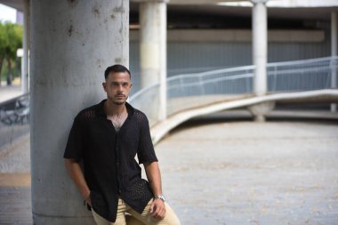 Genç, esmer, İspanyol, siyah tığ işi gömlekli modern bir adam, kameraya ciddi bir jestle bakıyor ve cebinde bir sütuna yaslanıyor. Moda, moda, gündelik konsept.