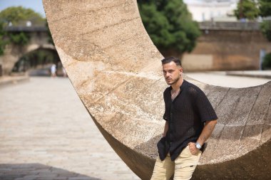 Genç, esmer, İspanyol, modern bir adam, siyah örgü örgü gömlek ve kot pantolon giyiyor, ciddi ve keskin bakışları var, elleri ceplerinde ve taş bir duvara yaslanıyor. Moda, gündelik konsept.