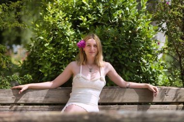 Genç, güzel, sarışın, beyaz elbiseli, kulağında menekşe çiçekleri olan, gülümseyen ve mutlu, açık hava parkındaki ahşap bankta oturan bir kadın. Güzelliği kavra, gençliği, milenyumu.