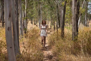 Genç bir kadın, güzel, sarışın, çiçekli bir elbise giyiyor, rahat ve sakin, ağaçların arasında yürüyor, doğa ve ekoloji ile bağlantılı, açık hava parkında. Güzellik kavramı, gençlik, milenyum.
