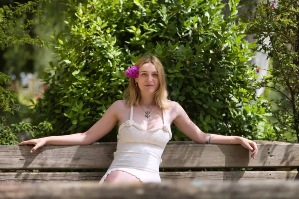 Genç, güzel, sarışın, beyaz elbiseli, kulağında menekşe çiçekleri olan, gülümseyen ve mutlu, açık hava parkındaki ahşap bankta oturan bir kadın. Güzelliği kavra, gençliği, milenyumu.