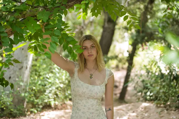 Genç, güzel, sarışın bir kadın, çiçekli bir elbise giyiyor, tatlı ve şefkatli bir bakışla, bir ağacın dalına tutunuyor, doğa ve ekolojiyle bağlantılı, açık hava parkında. Güzellik kavramı, gençlik.