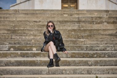 Genç, güzel, sarışın, deri ceketli, yılan etekli, çizmeli ve güneş gözlüklü, rahat ve sakin bir şekilde merdivende oturan kameraya bakıyor. İsyankar, boyun eğmez bir kavram..