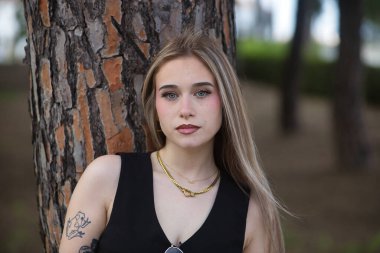 Genç, güzel bir kadının portresi, sarışın, yeşil gözlü, siyah bluzlu, bir ağacın gövdesine yaslanırken kameraya tatlı ve nazikçe bakan. Masumiyet kavramı.