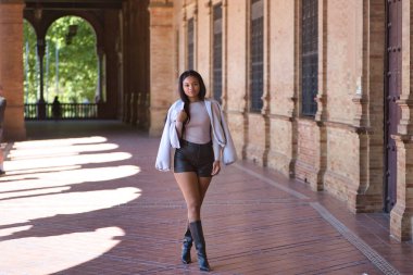 Genç, güzel, Afrikalı bir kadın, ceketli, deri şortlu ve botlu, İspanya meydanında poz veriyor ve modellik yapıyor. Moda konsepti, güzellik, moda.