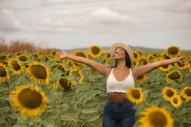 Genç, güzel, Latin esmer kadın beyaz örgü örer, kot pantolon giyer ve şapka giyer, kolları açık, minnettar ve mutlu bir şekilde ayçiçeği tarlasının ortasında. Kavram: doğa, tarım.