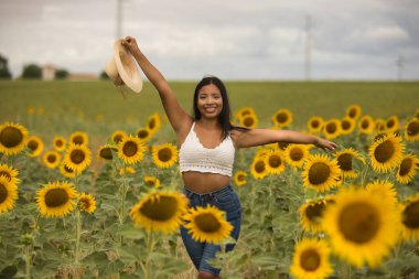 Genç, çekici, latin esmer kadın beyaz örgü örer, kot pantolon giyer ve elinde bir şapka, kolları ayçiçeği tarlasında mutlu bir şekilde gülümserdi. Kavram: doğa, tarım.