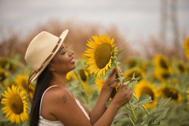 Genç, güzel, latin esmer kadın beyaz örgü örer ve şapka takar, ayçiçeği tarlasının ortasında bir bitkiyi tutarken mutlu bir şekilde gülümser. Kavram: doğa, tarım, kırsal.